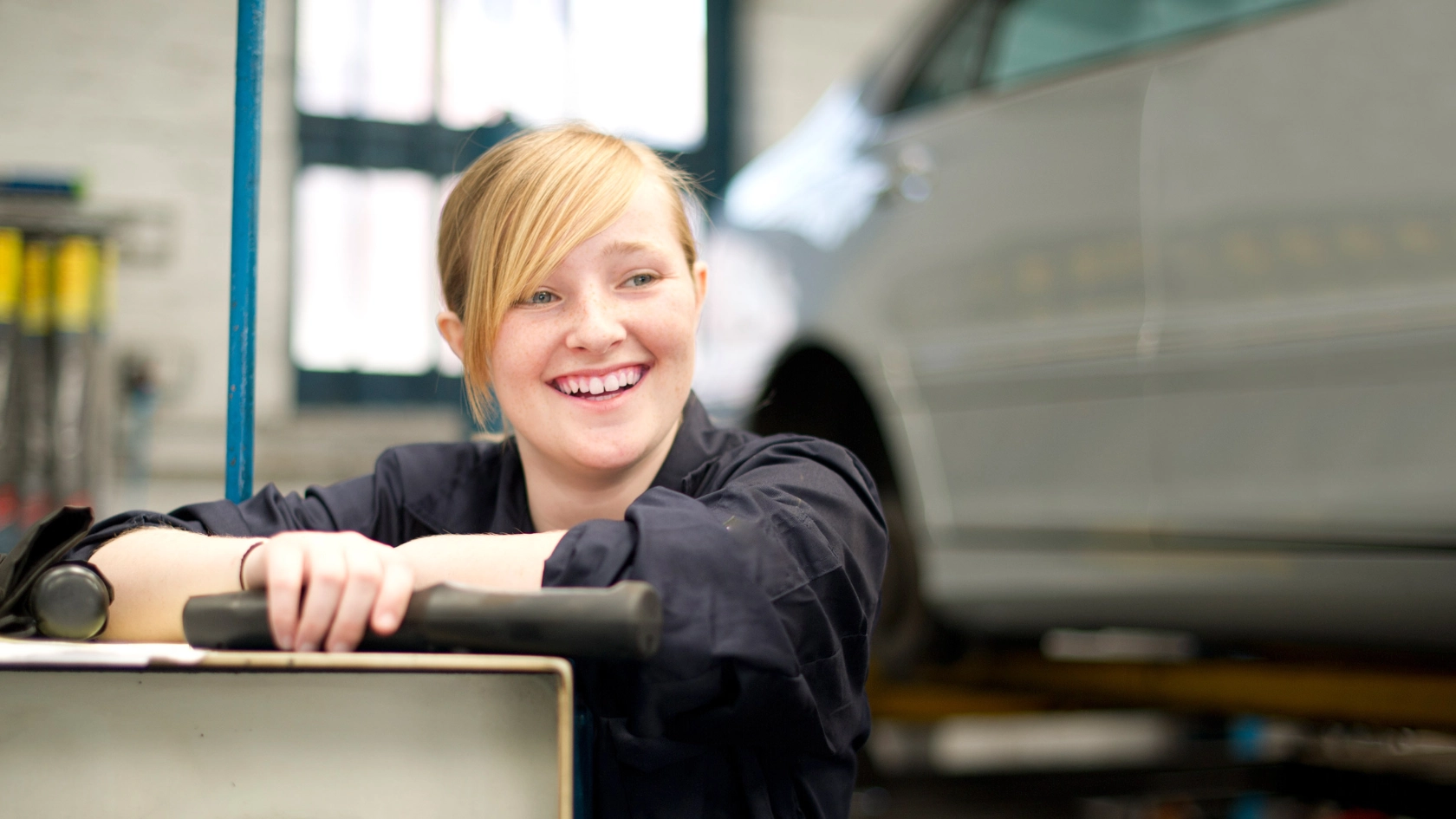 Eine junge blonde Frau mit einem Werkzeug in der Hand steht lächelnd in einer KFZ-Werkstatt.