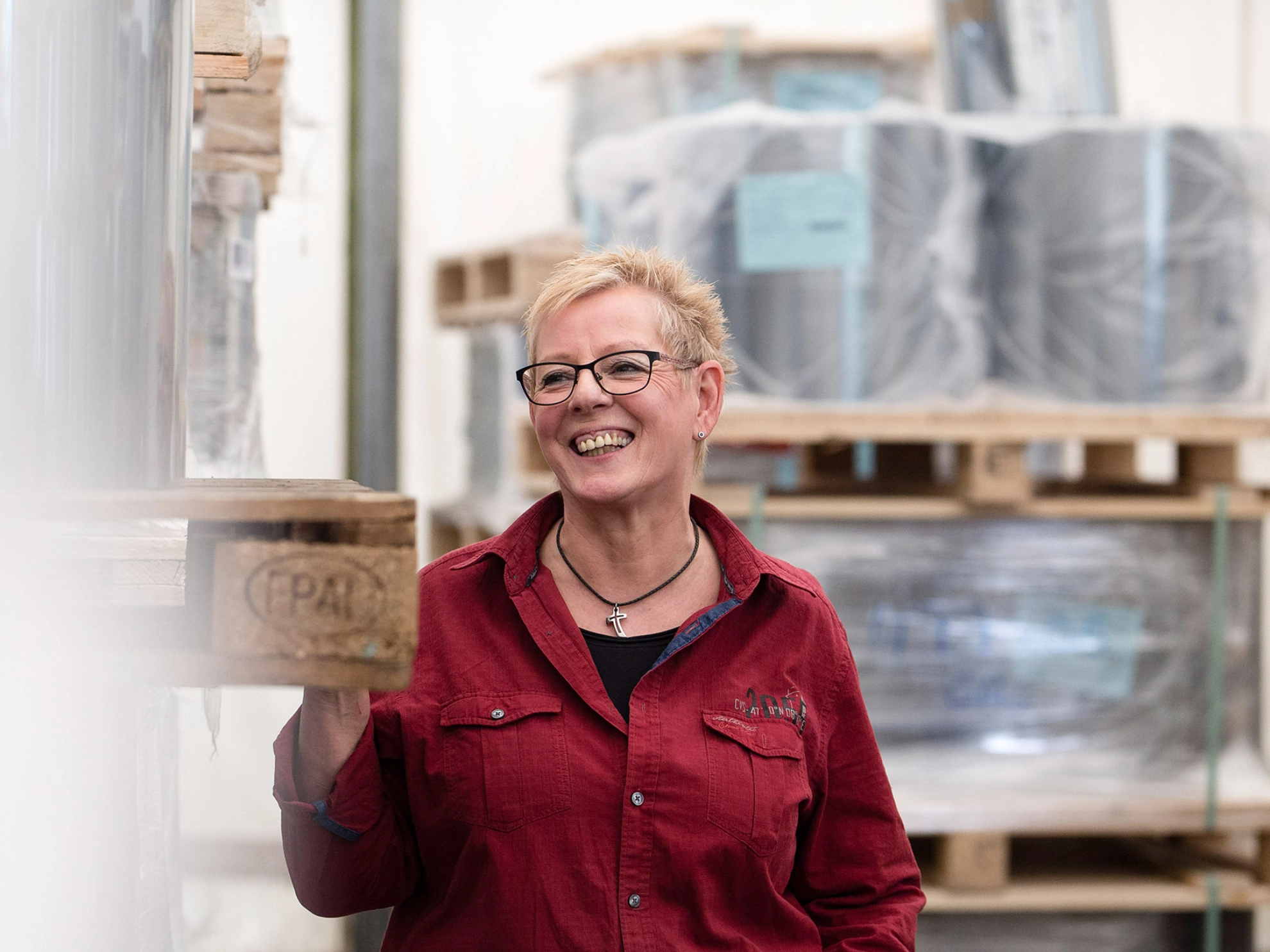 Eine Frau in einem dunkelroten Hemd steht lachend neben einer Palette mit Plastikfolie.