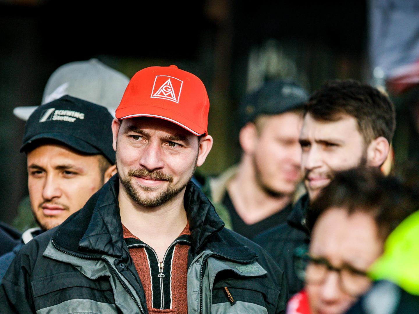 Ein junger Mann mit IG Metall-Kappe steht in eine Gruppe Menschen und lächelt.
